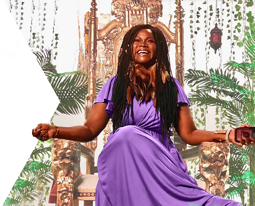 Abiola Abrams with a big smile sitting on a golden throne with palm fronds in the background